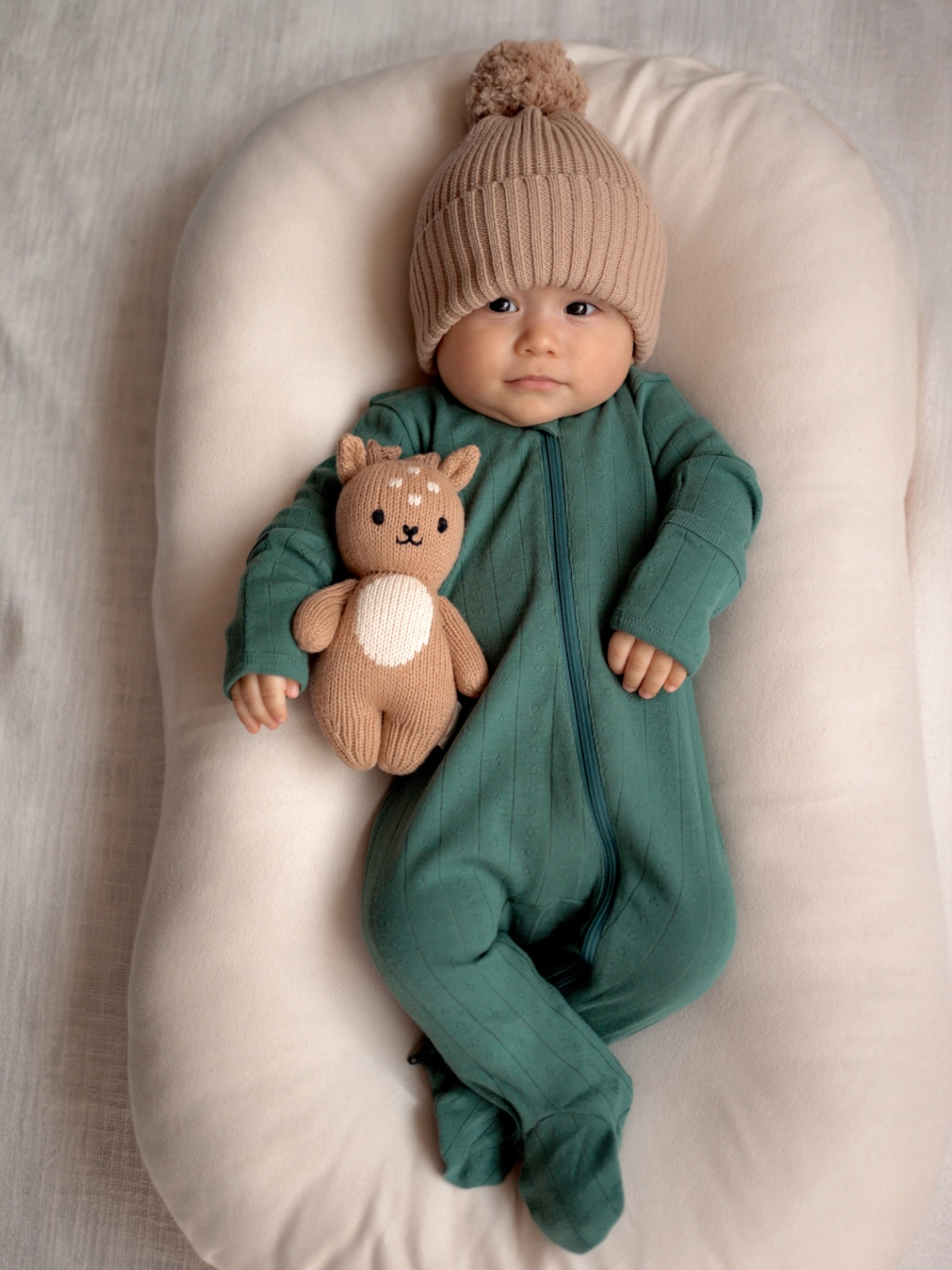 Infant in green onesie and knitted hat, holding a plush toy, lying on a soft, cream-colored surface.