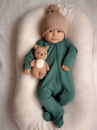 Infant in green onesie and knitted hat, holding a plush toy, lying on a soft, cream-colored surface.