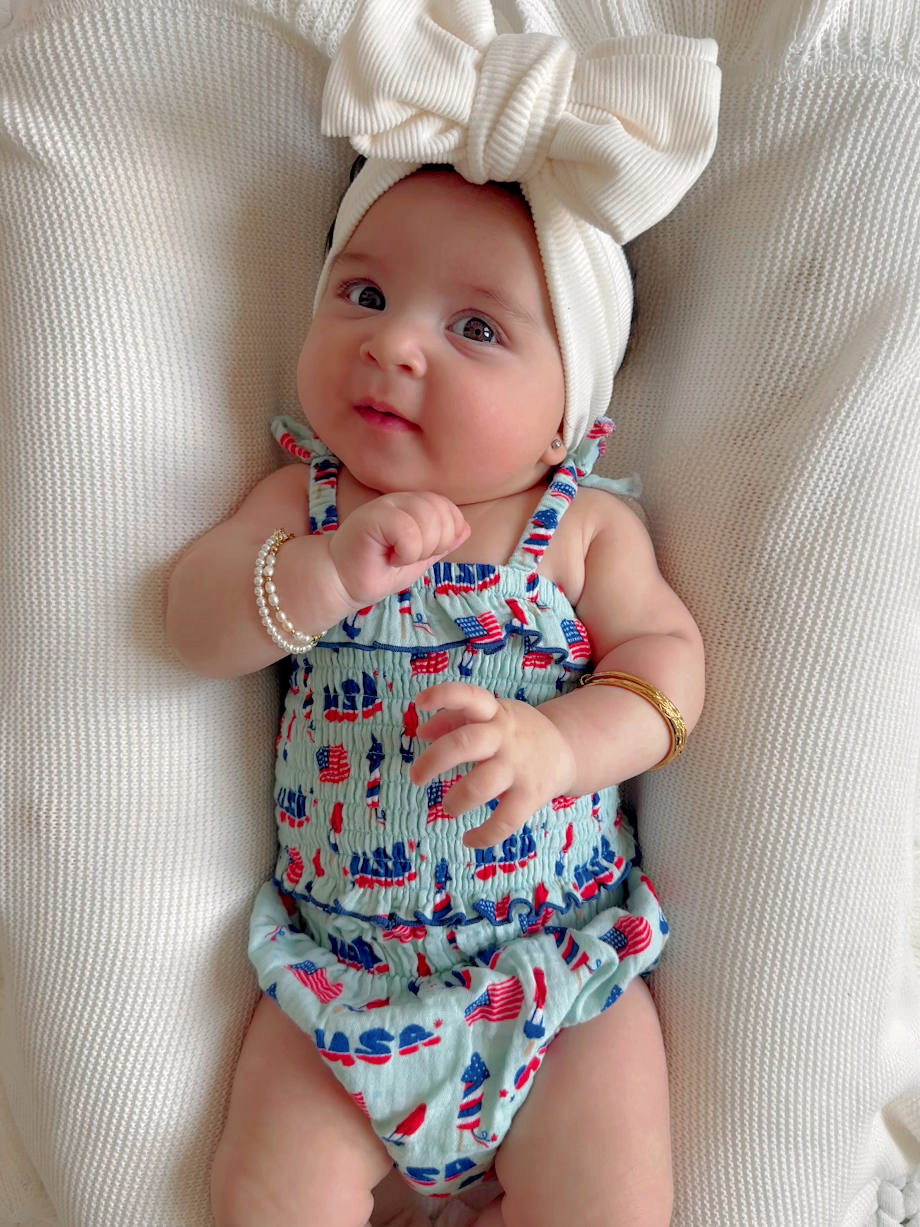 Baby girl in a cute swimsuit with patriotic patterns, wearing a large bow and playful expression, sitting on soft fabric.