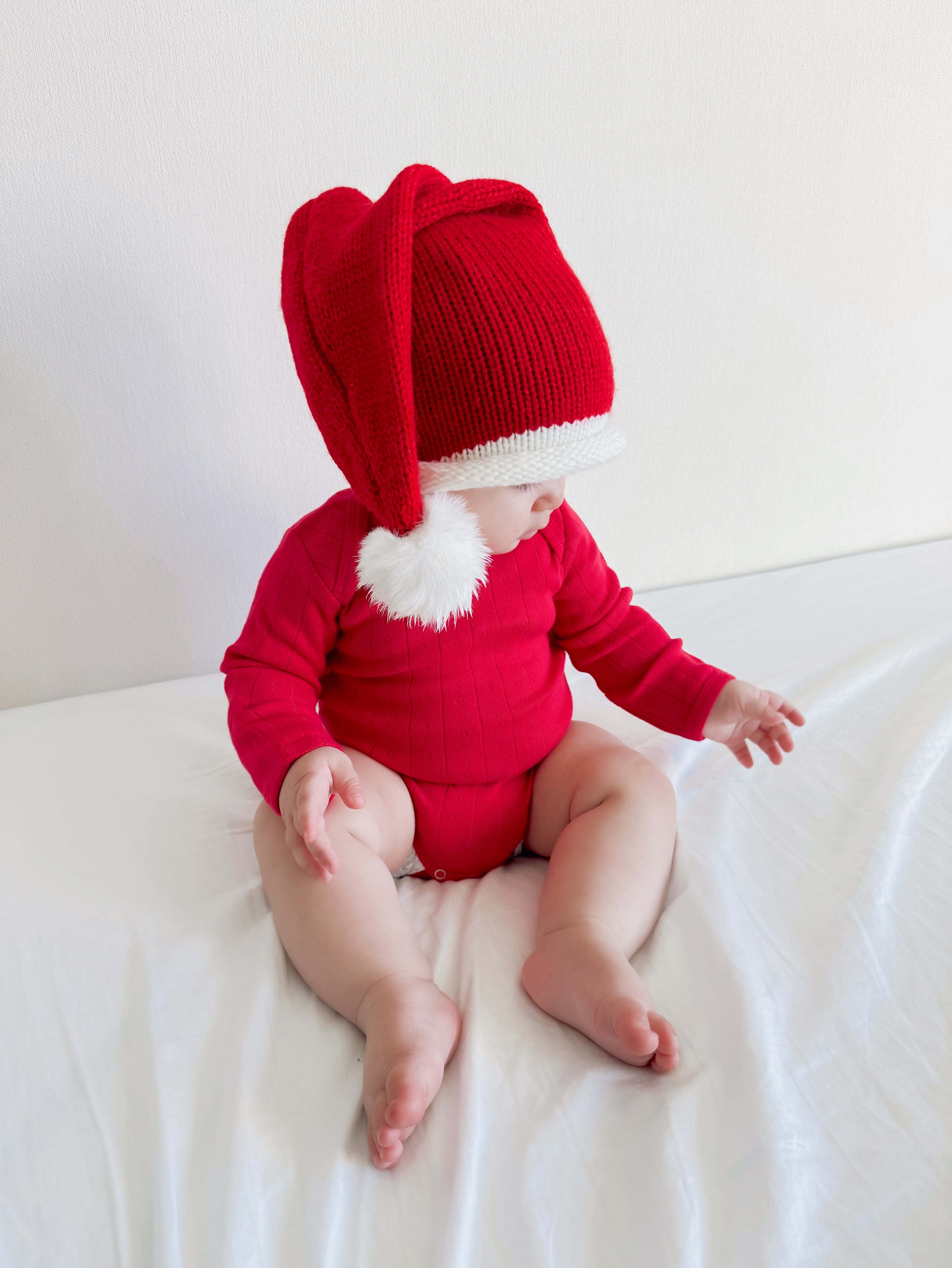 Baby wearing a red outfit and a festive Santa hat, sitting on a white bed, smiling with hands on knees.