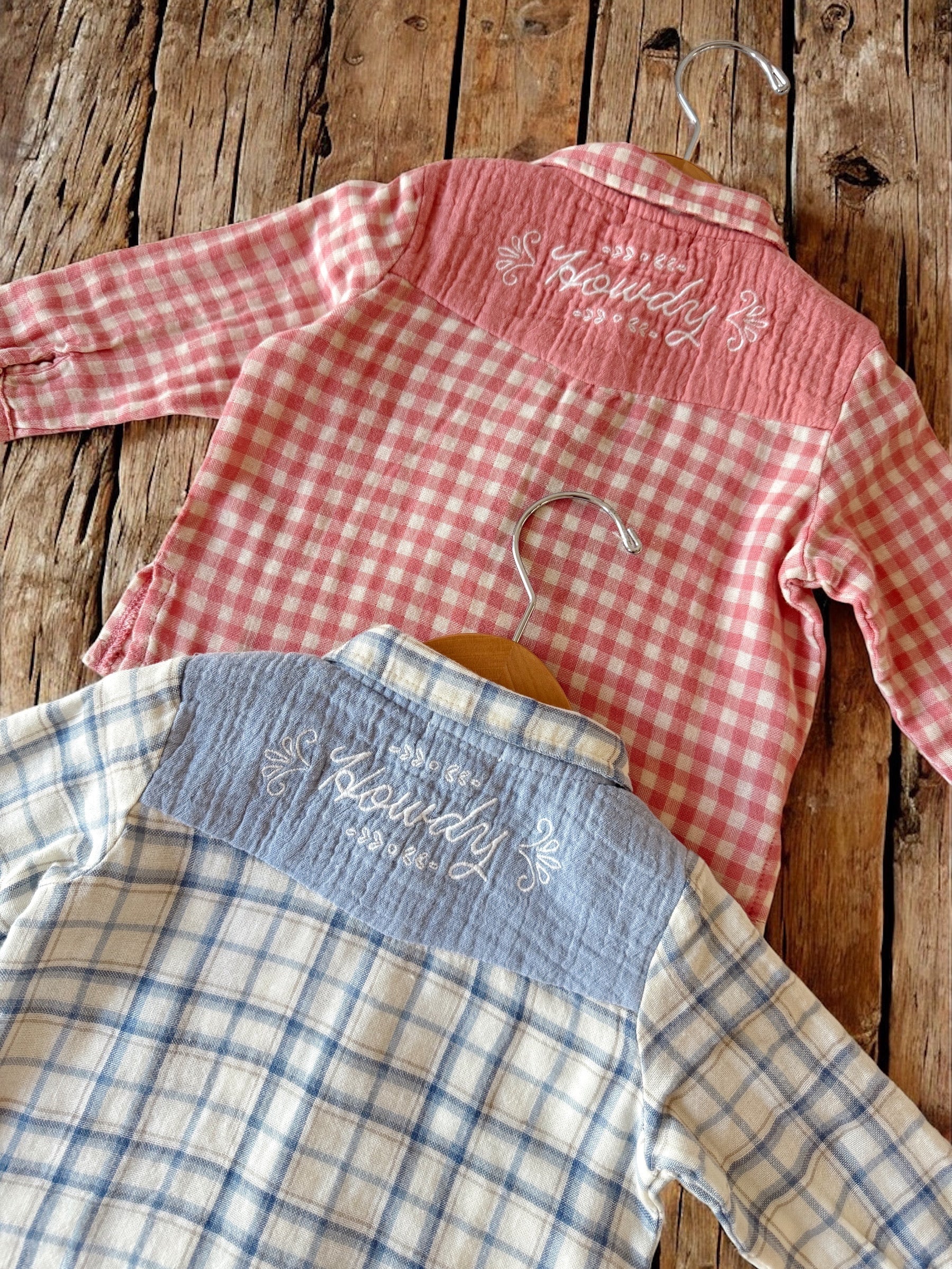 Two plaid shirts hanging on hooks, one pink and one blue, both featuring "Howdy" embroidery on the back.