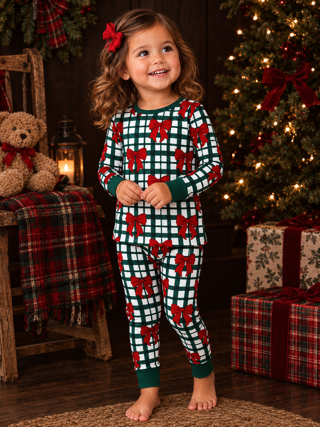 Smiling girl in festive pajamas with red bows, standing by Christmas tree and teddy bear decorations.