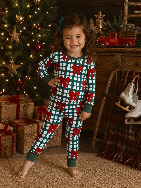 Smiling girl in holiday pajamas with red bows, standing near a Christmas tree and wrapped gifts.