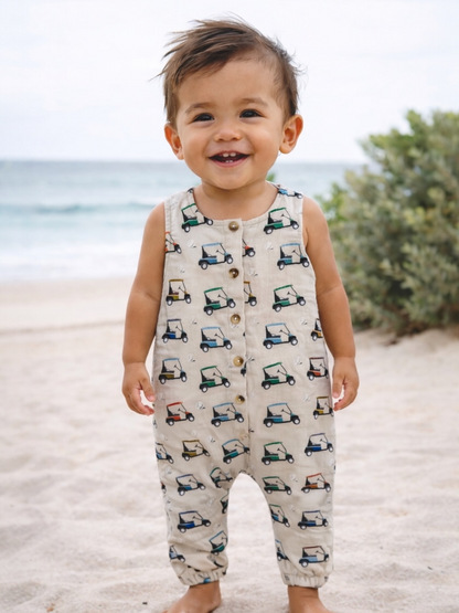 Smiling toddler in a patterned romper stands on the beach, with ocean waves and greenery in the background.