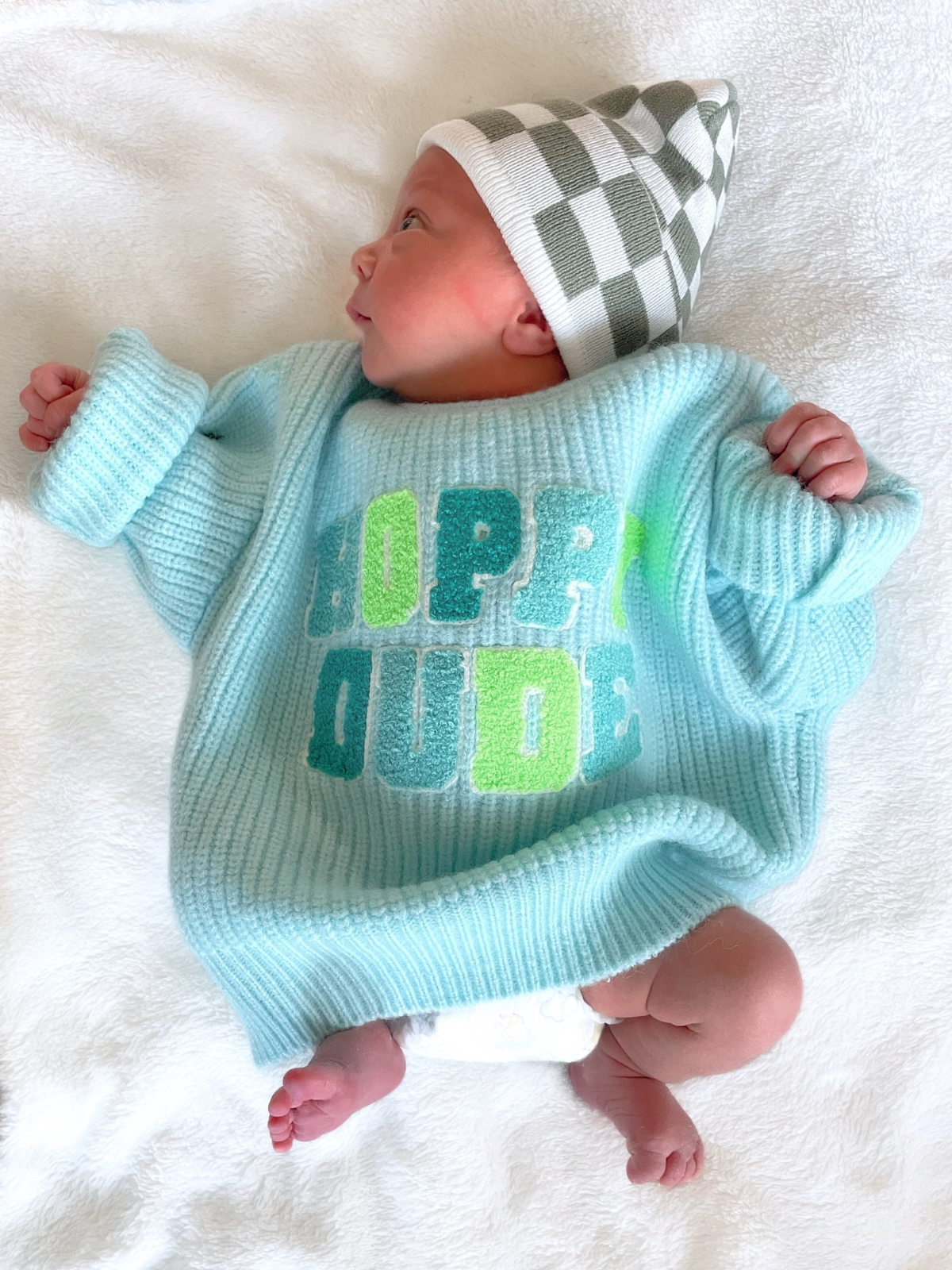 Baby in blue sweater with "HOPPER DUDE" text and checkered hat, lying on a soft white blanket.