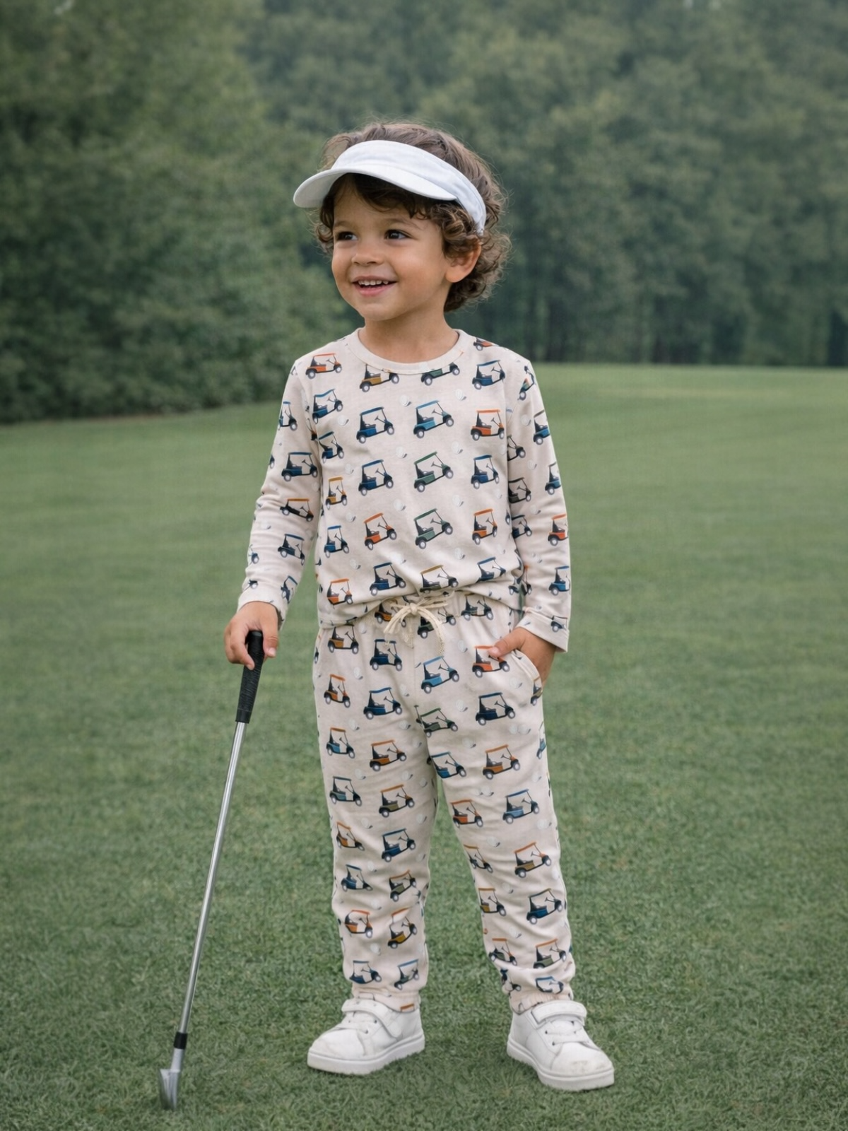 Child in golf-themed pajamas and a visor, smiling on a green field with trees in the background.