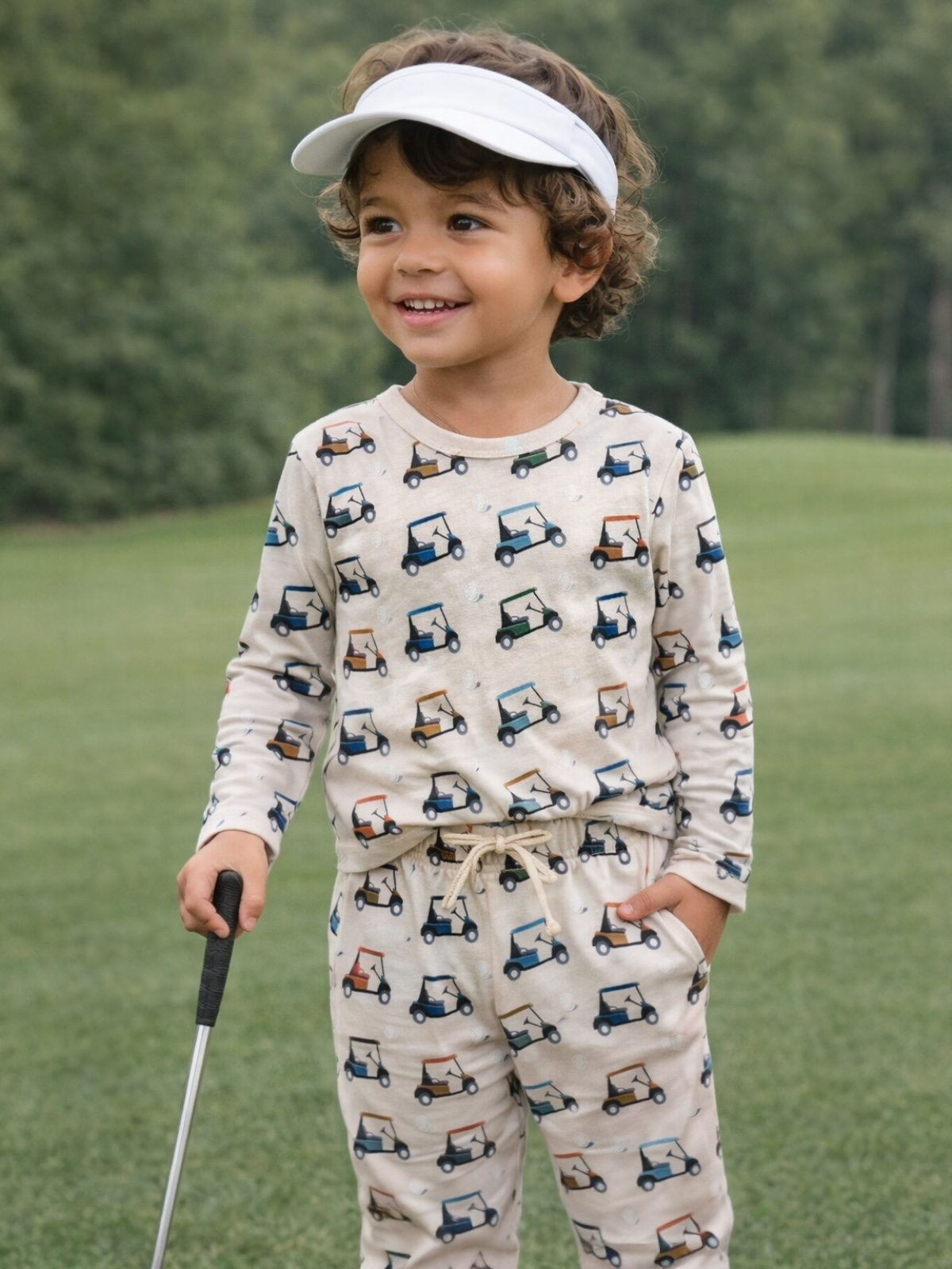 Smiling child in golf-themed pajamas, standing on a green field, holding a golf club, wearing a visor.