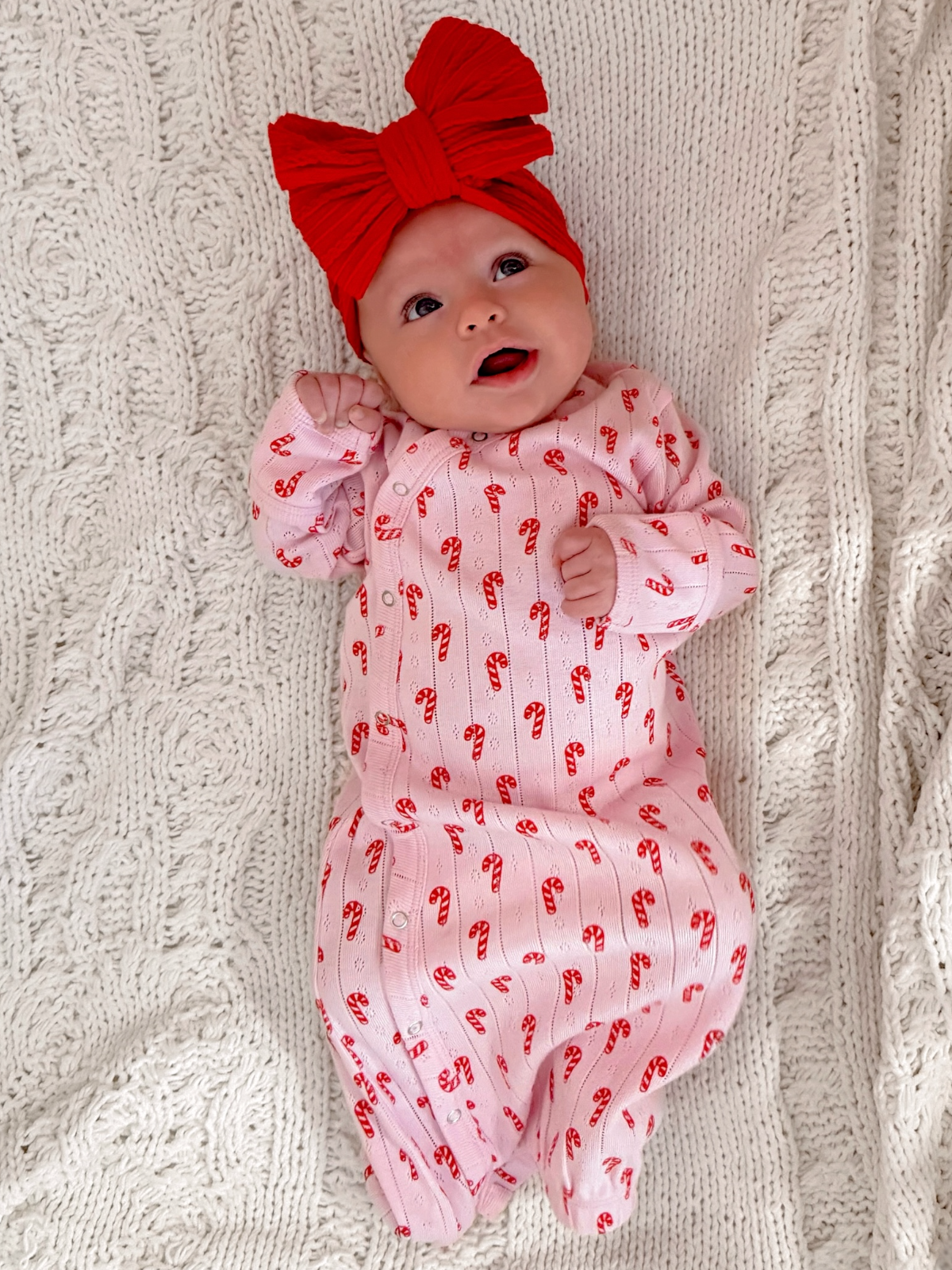 Smiling baby in candy cane pajamas and a red bow, lying on a textured white blanket.