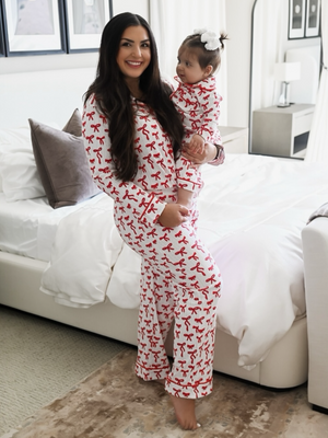 Smiling woman in matching red ribbon pajamas holds a baby in similar outfit, standing in a cozy bedroom.