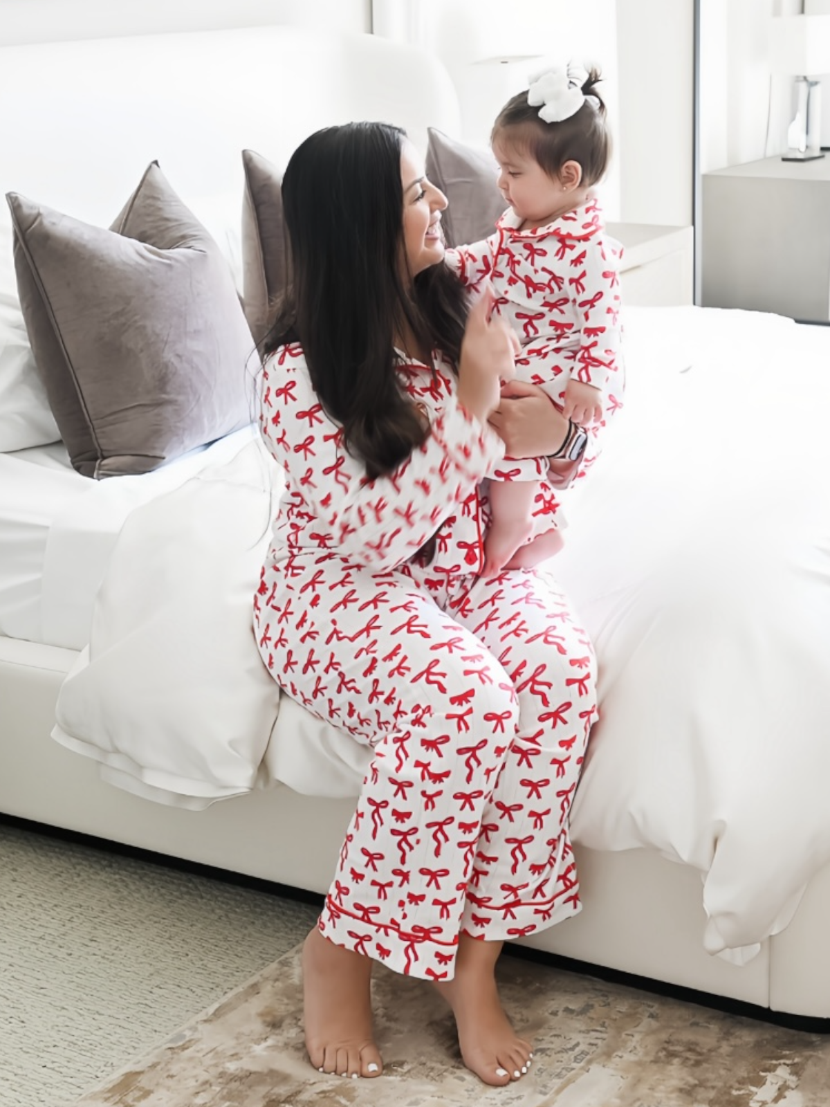 Woman and child in matching red ribbon pajamas, sharing a joyful moment on a bed with neutral tones.