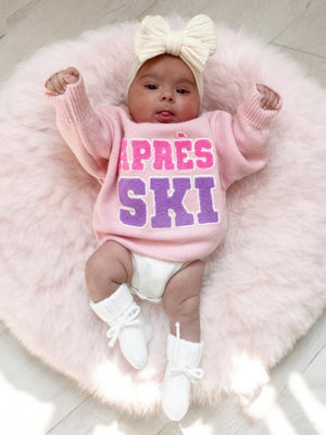 Baby in pink "Après Ski" sweater, lying on a fluffy pink rug, wearing a bow headband and white booties.