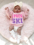 Baby in pink "Après Ski" sweater, lying on a fluffy pink rug, wearing a bow headband and white booties.
