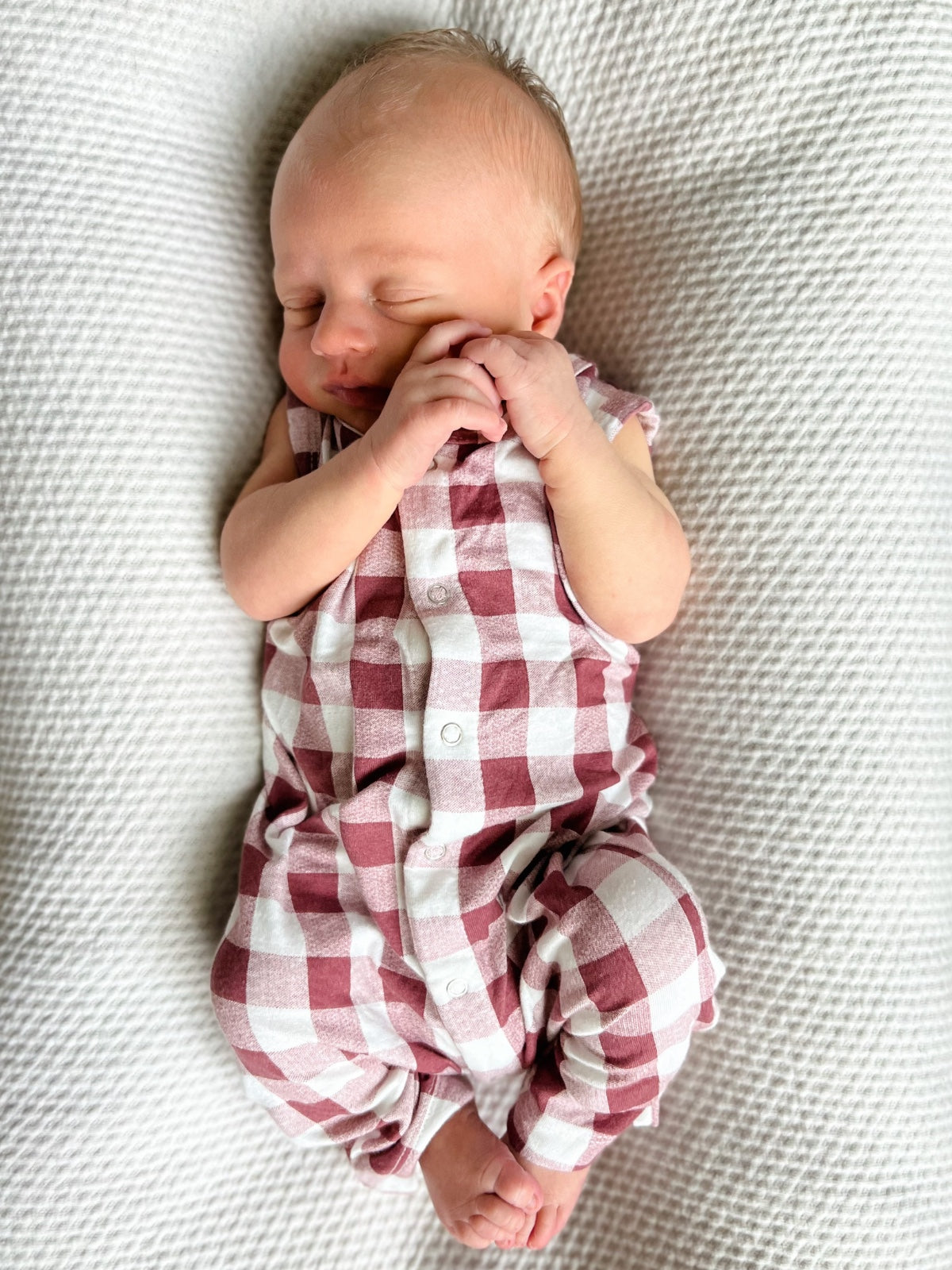 Newborn baby peacefully sleeping in a red and white checkered outfit on a textured blanket.