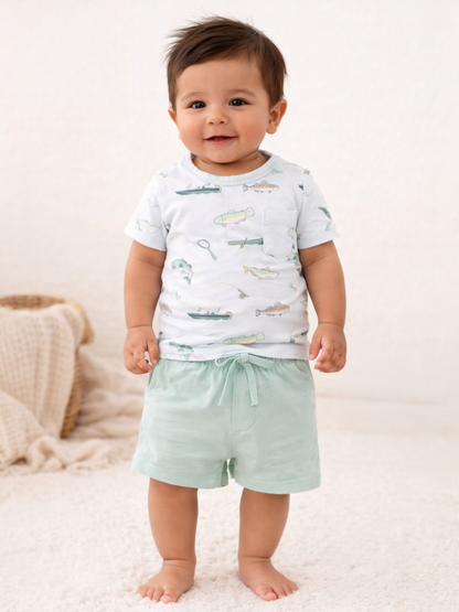 Smiling toddler wearing a fish-patterned shirt and mint shorts, standing on a soft white rug.