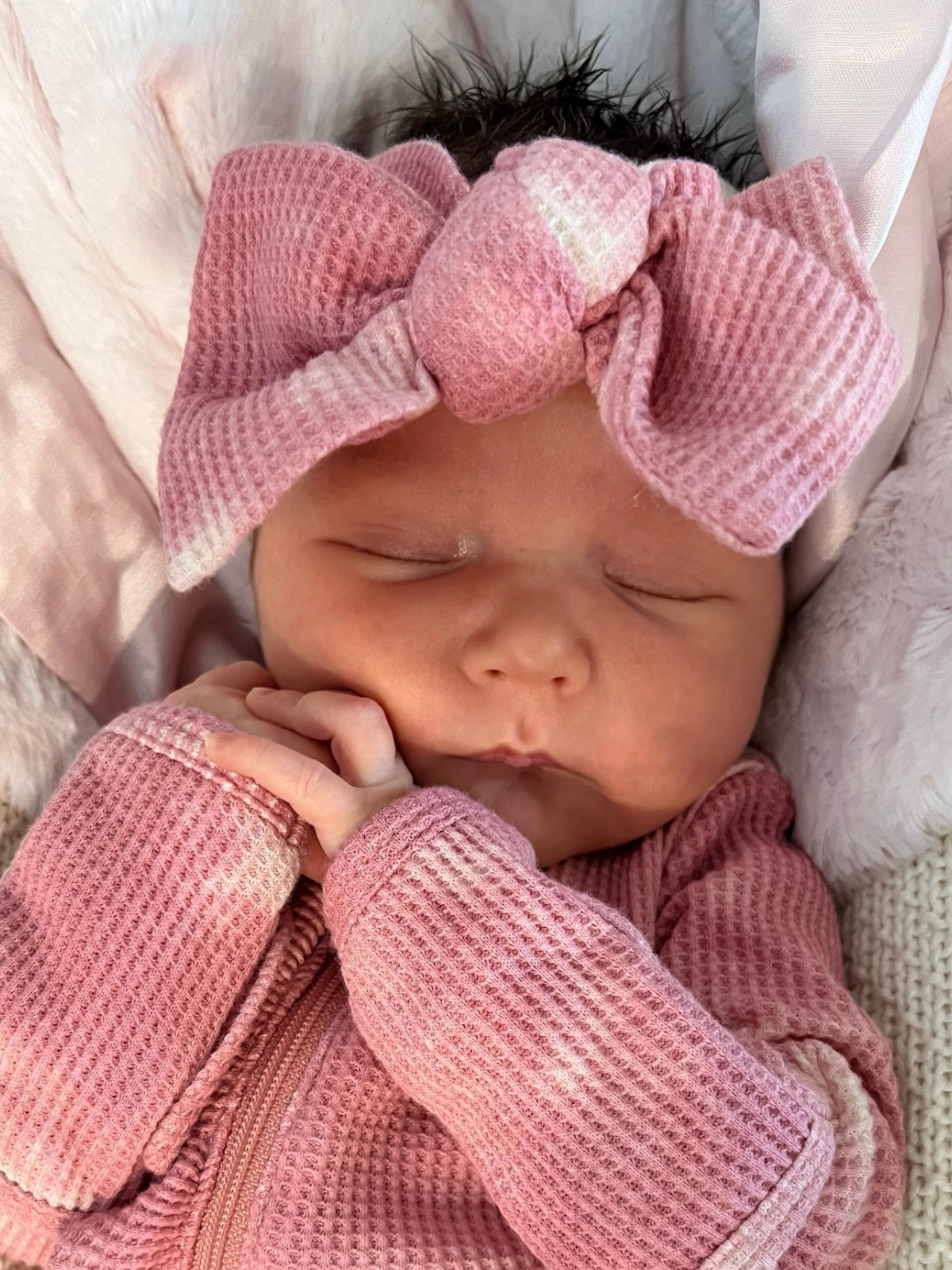 Newborn baby girl sleeping peacefully in pink outfit with a large bow, resting on a soft blanket.