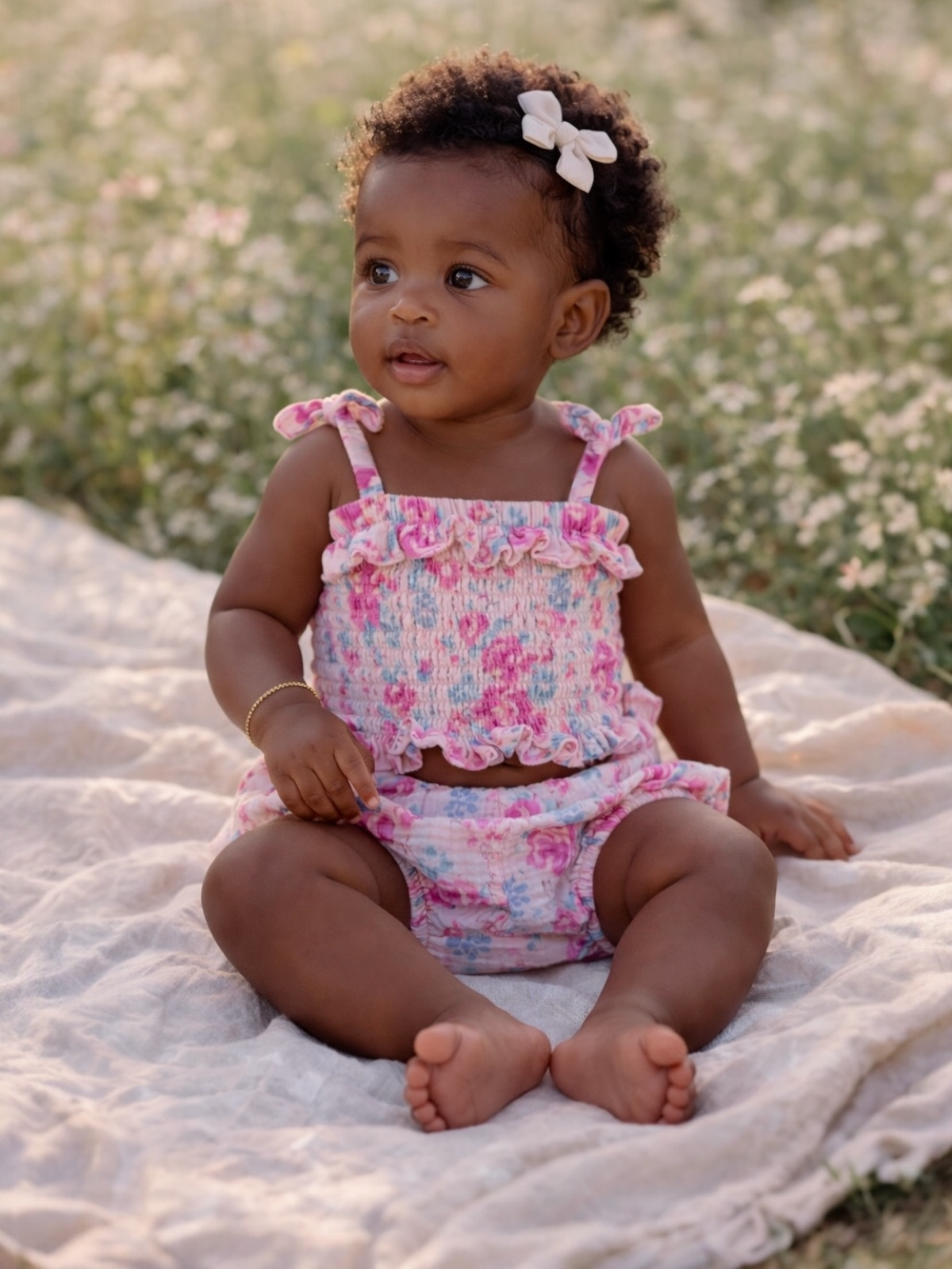 Baby girl in floral outfit sits on a blanket in a field of flowers, smiling and looking to the side.