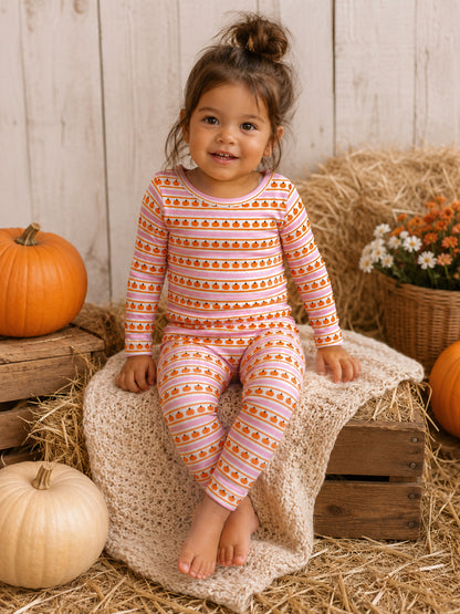 Smiling child in pumpkin-themed pajamas, sitting on straw and a cozy blanket with pumpkins and flowers around.