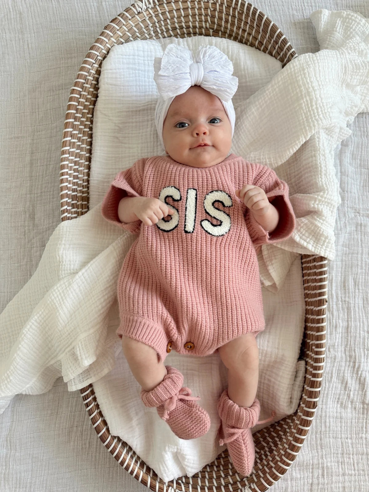Infant in a pink knitted romper with "SIS" text, wearing a white bow headband, resting in a woven basket.