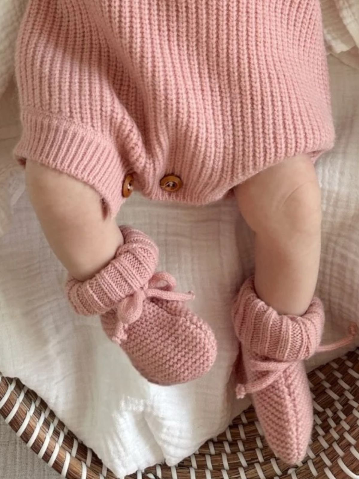 Baby's legs wearing pink knitted booties, resting on a soft blanket in a woven basket.