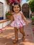 Smiling toddler in floral dress and sandals, walking on a sunny path with vibrant flowers and a wooden door behind.