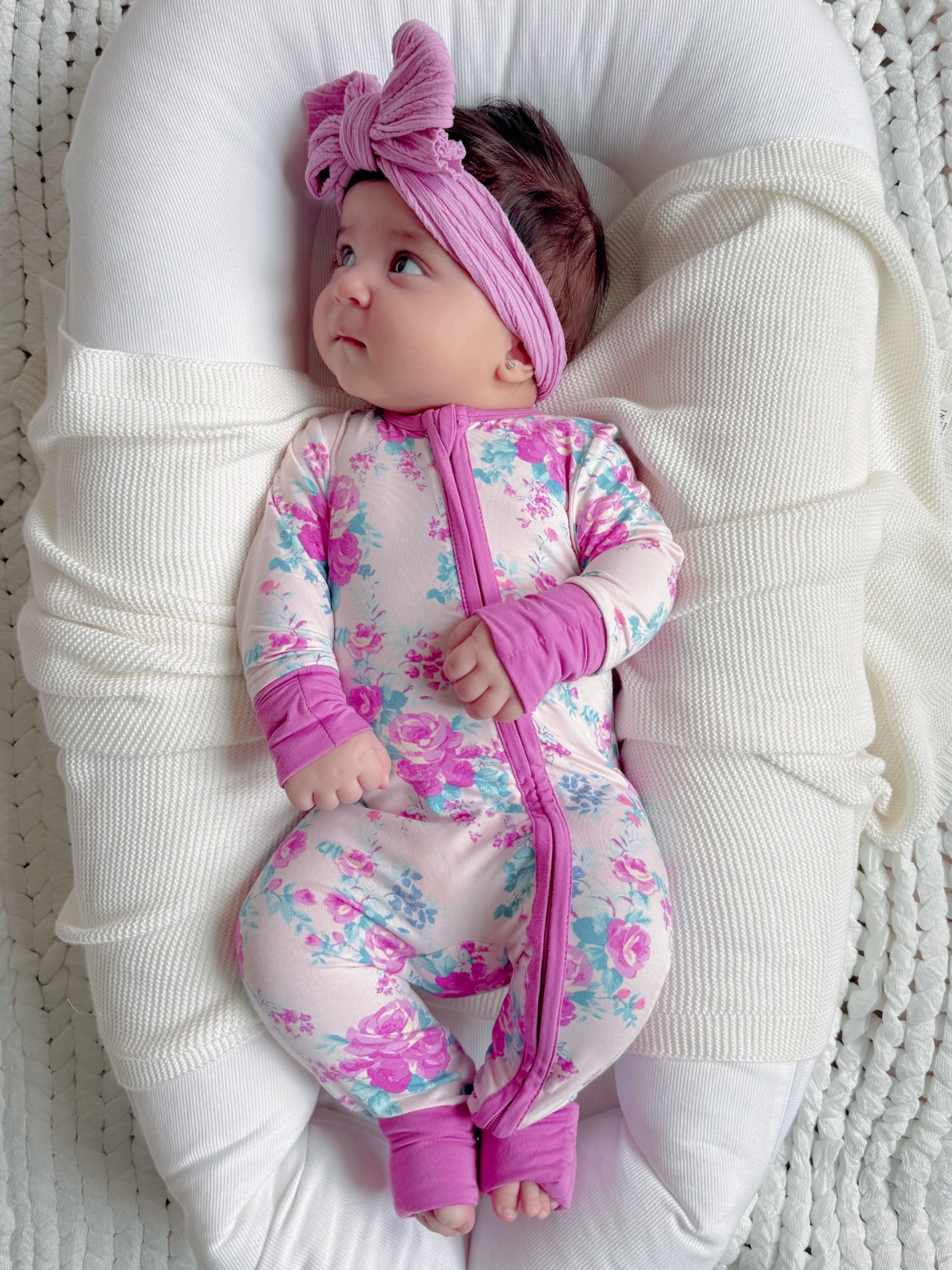 A baby in a floral outfit and pink headband lies on a cozy blanket, gazing curiously.