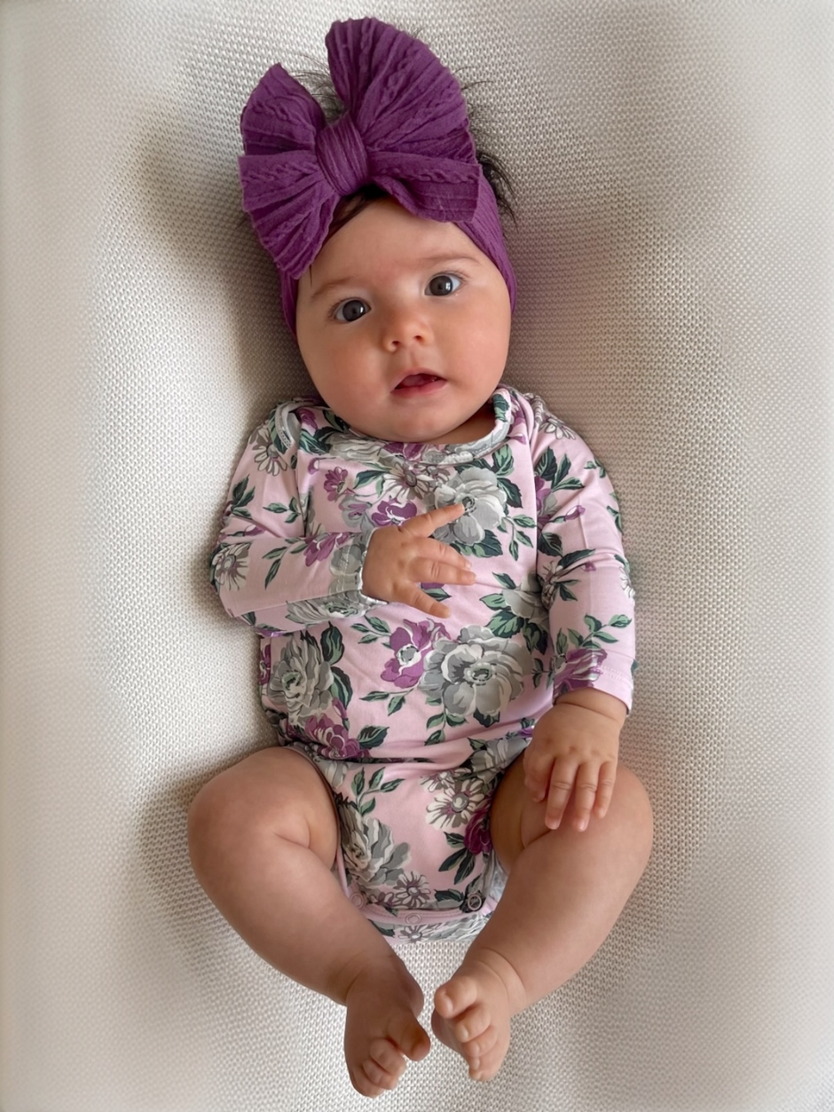 Infant girl lying on a blanket, wearing a floral outfit and a large purple headband, with a curious expression.