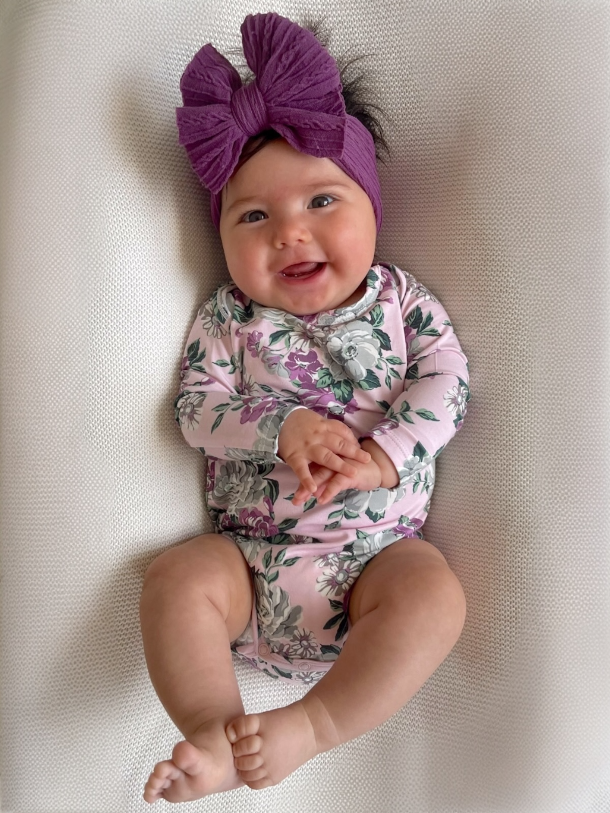 Smiling baby girl in a floral onesie and purple headband, resting on a textured surface.