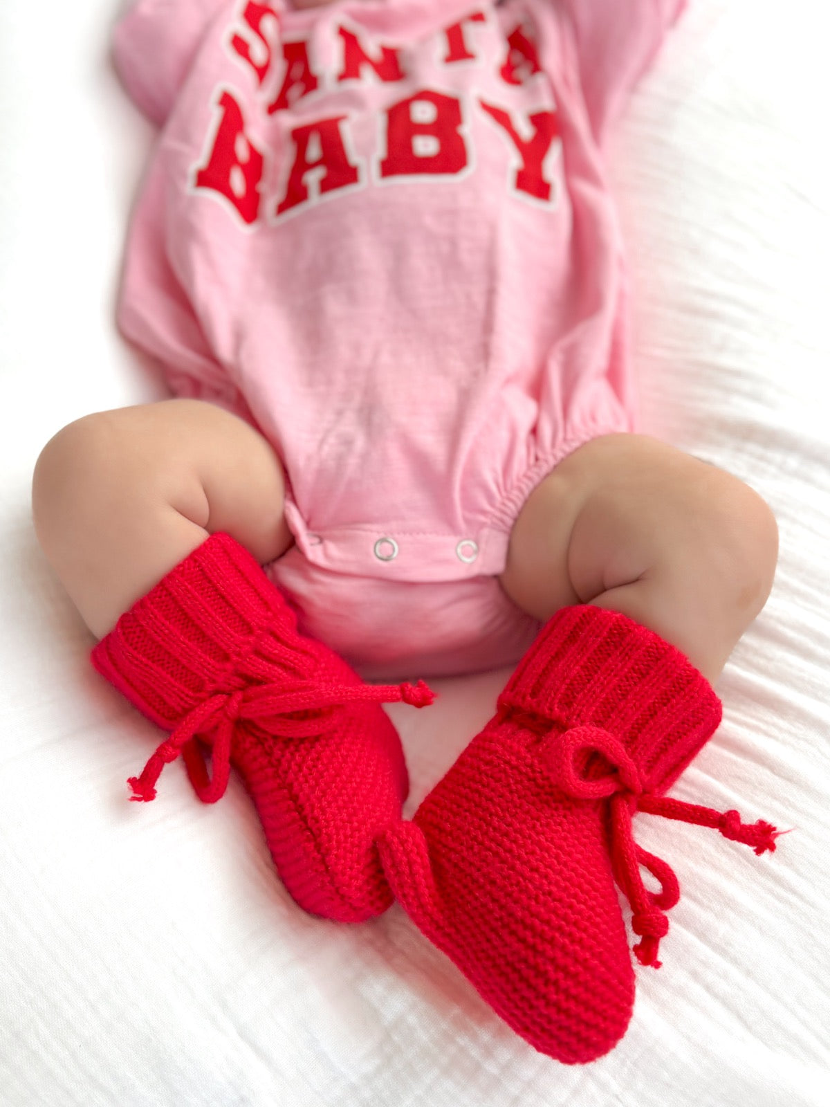 Baby wearing a pink "Santa Baby" onesie with cozy red booties on a soft white blanket.