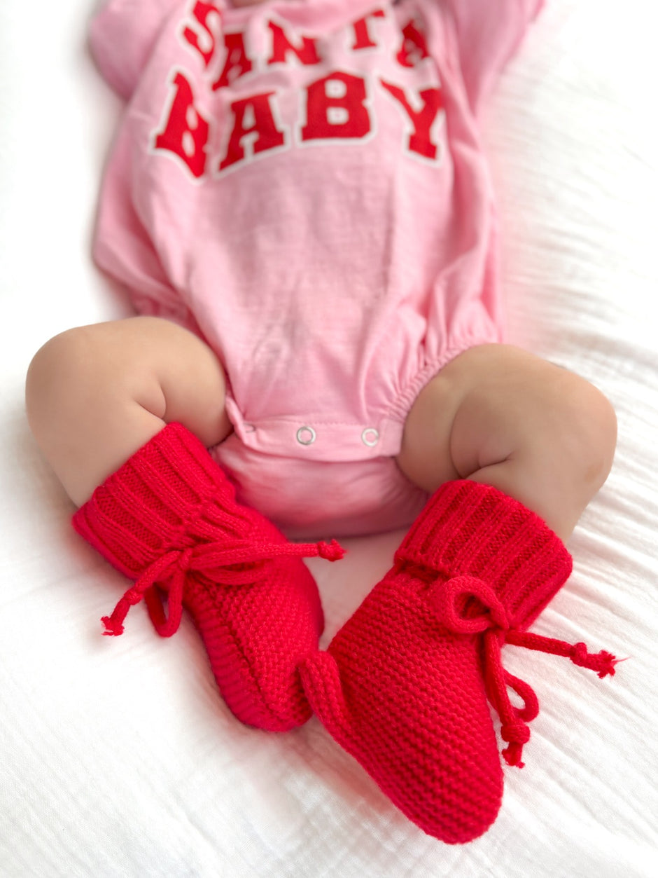 Baby wearing a pink "Santa Baby" onesie with cozy red booties on a soft white blanket.