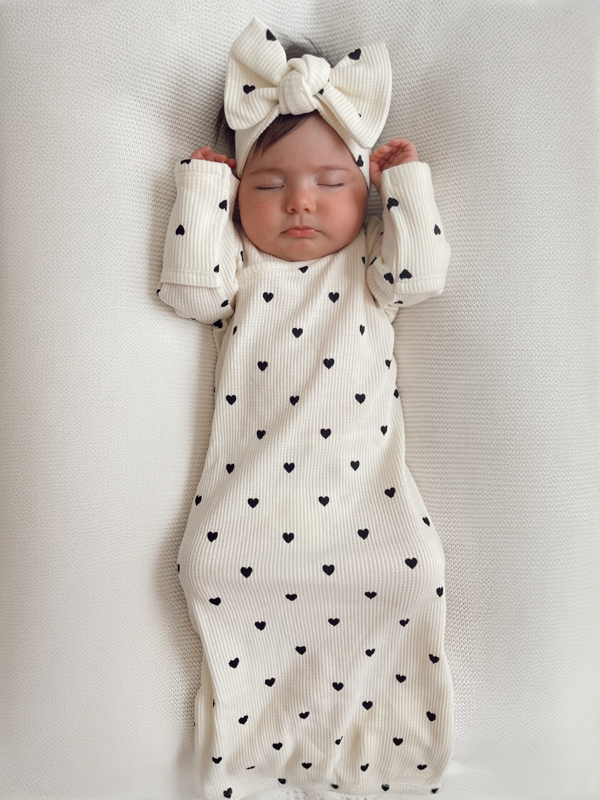 Baby girl sleeping peacefully in a cream outfit with black hearts and a matching headband. Soft textured background.