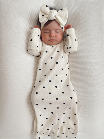 Baby girl sleeping peacefully in a cream outfit with black hearts and a matching headband. Soft textured background.