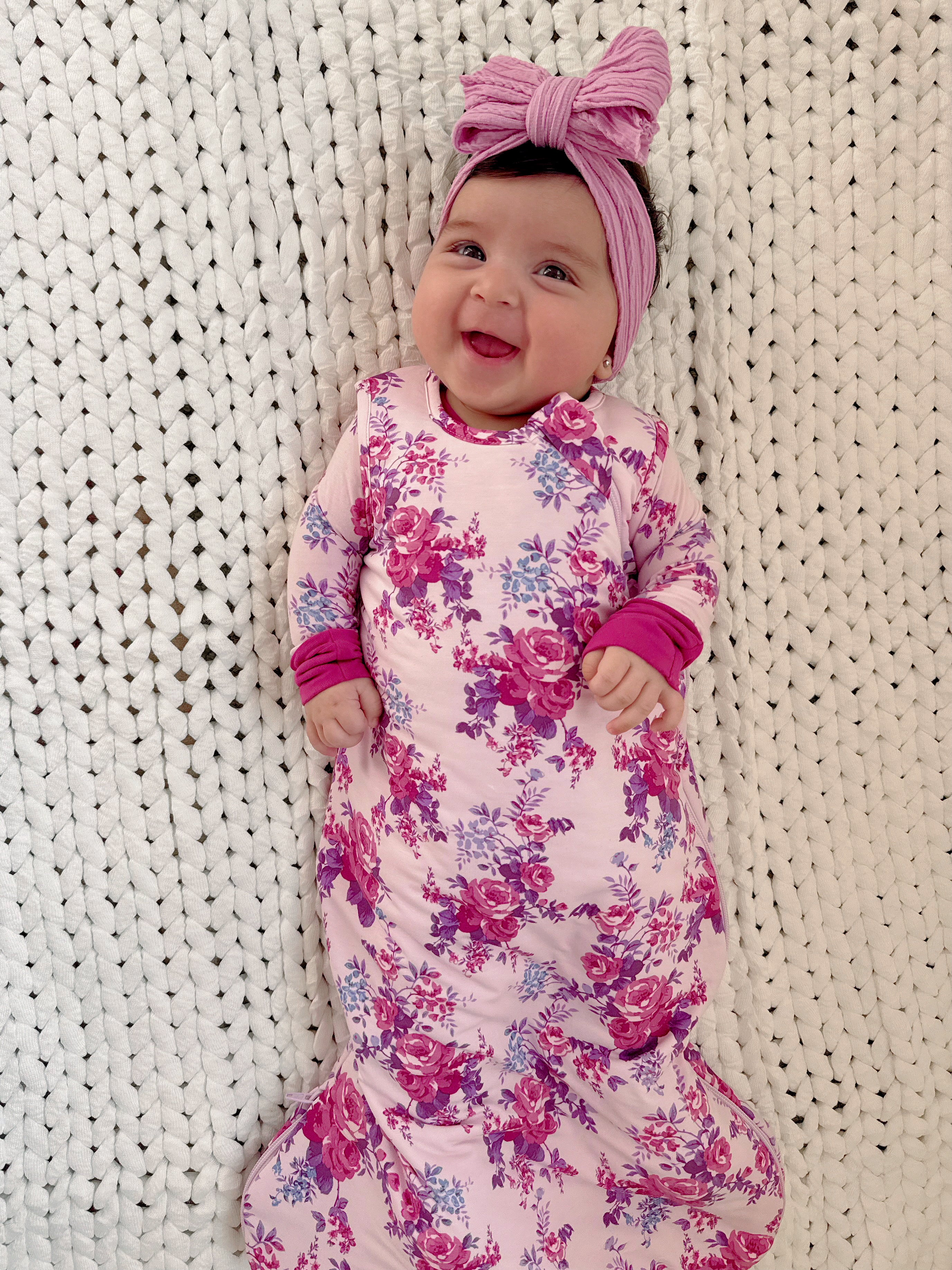Baby girl smiles while wearing a floral pink outfit and matching headband on a textured white blanket.