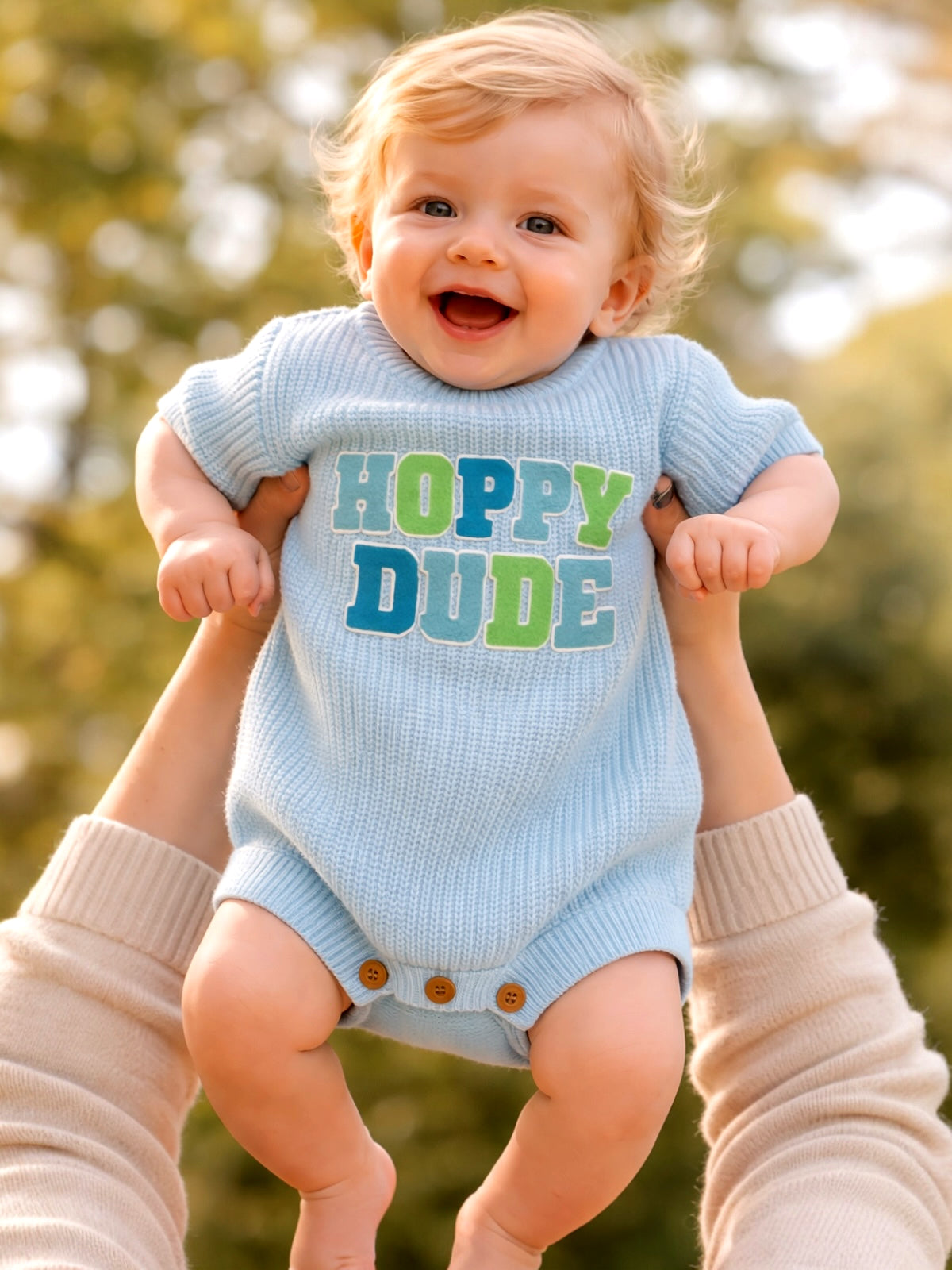 Smiling baby in a blue onesie with "Hoppy Dude" text, held by a person against a blurred outdoor background.