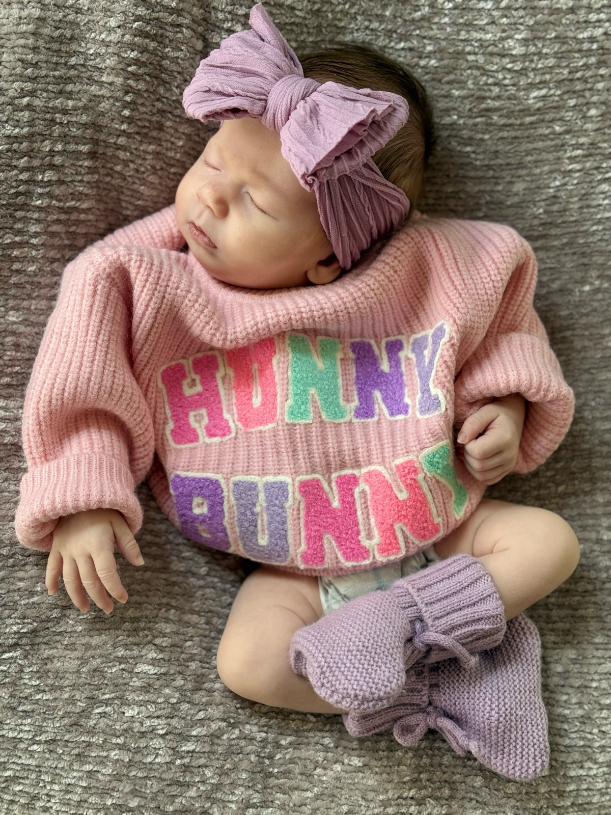 Baby girl in pink "Hunny Bunny" sweater and purple headband, resting on a cozy gray blanket.