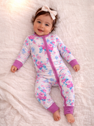 Smiling baby in floral pajamas and a bow, lying on a soft blanket with a playful expression.
