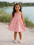Smiling girl in a pink bunny-patterned dress, standing by a lake with green trees in the background.