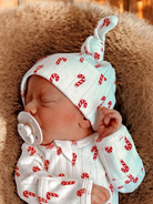Newborn asleep in candy-cane patterned outfit and hat, with a pacifier, on a soft texture background.