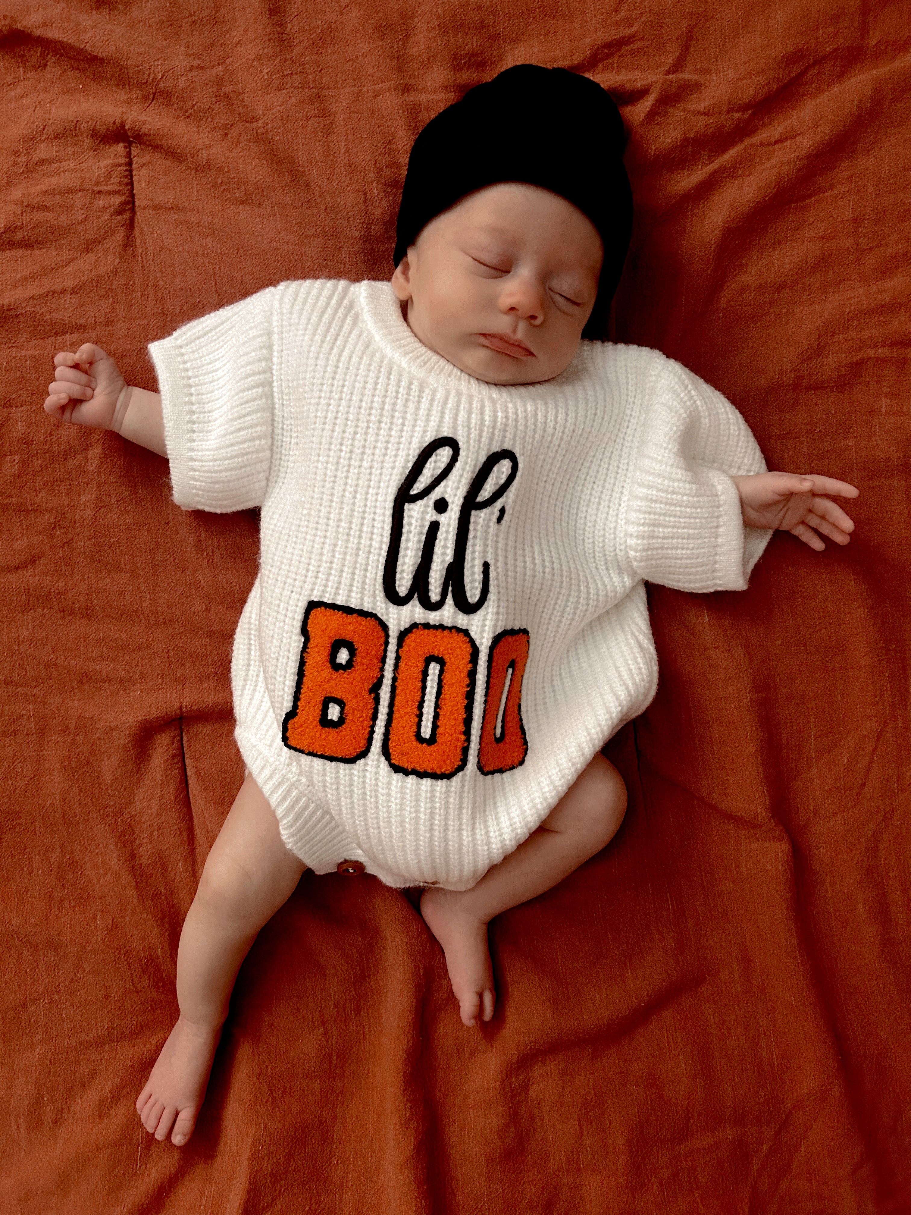 Infant wearing a white sweater with "lil' BOO" in orange, sleeping on an orange fabric background.