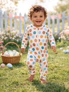 Smiling toddler in polka dot pajamas, standing on grass near easter eggs and flowers with a festive backdrop.
