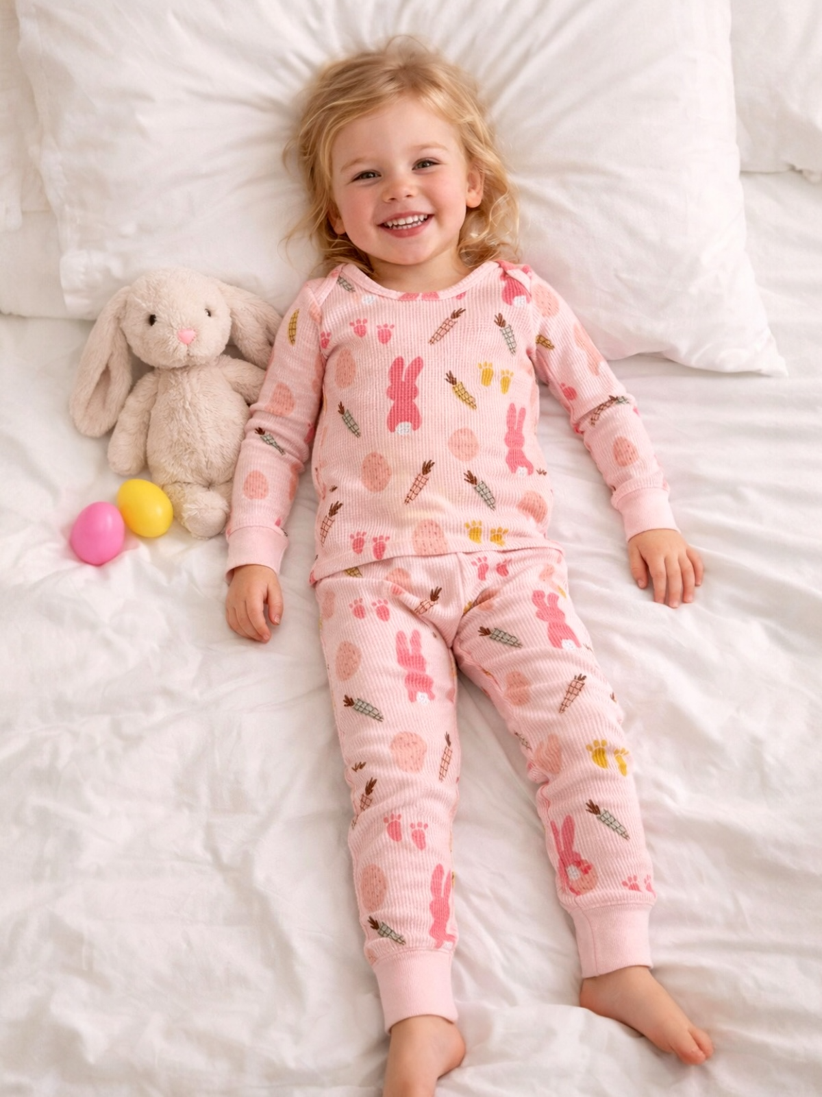 Smiling child in bunny-themed pajamas lying on a bed with a stuffed rabbit and colorful eggs.