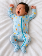 Baby in a blue onesie with bunny and vegetable patterns, yawning while lying on a soft white blanket.