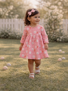 Smiling child in a pink bunny-patterned dress walks on grass, with a floral background and soft sunlight.
