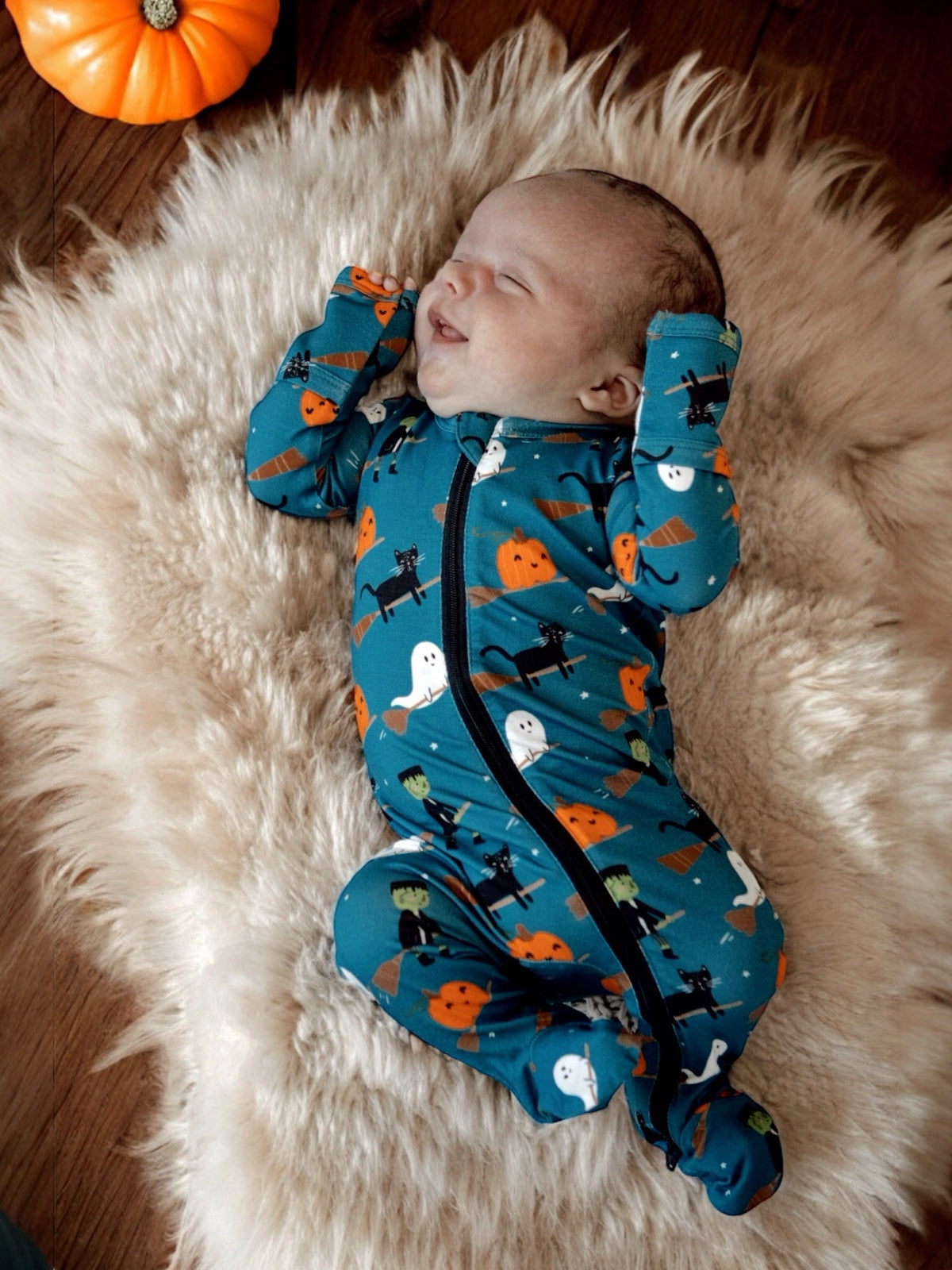 Baby smiling while lying on a furry blanket, dressed in a blue Halloween-themed onesie with pumpkins and ghosts.
