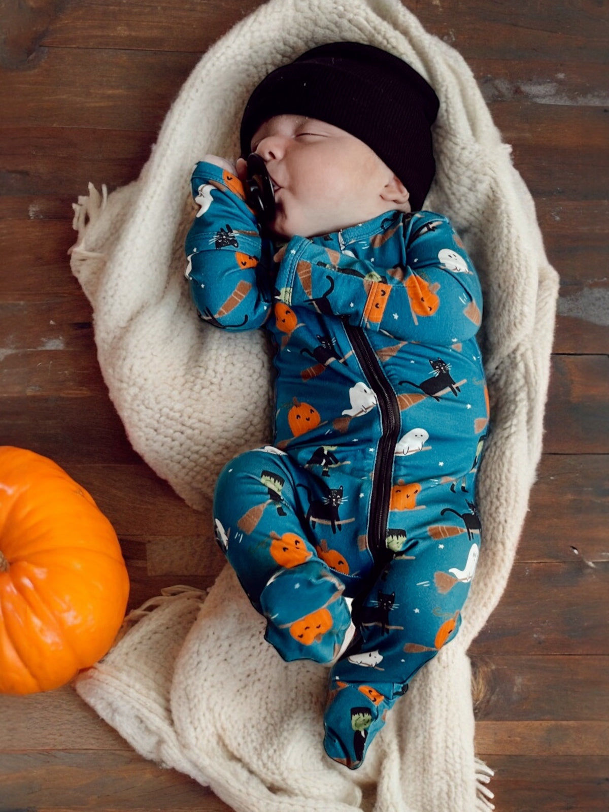 Baby sleeping on a cozy blanket, wearing a blue Halloween-themed onesie, with a pumpkin nearby.