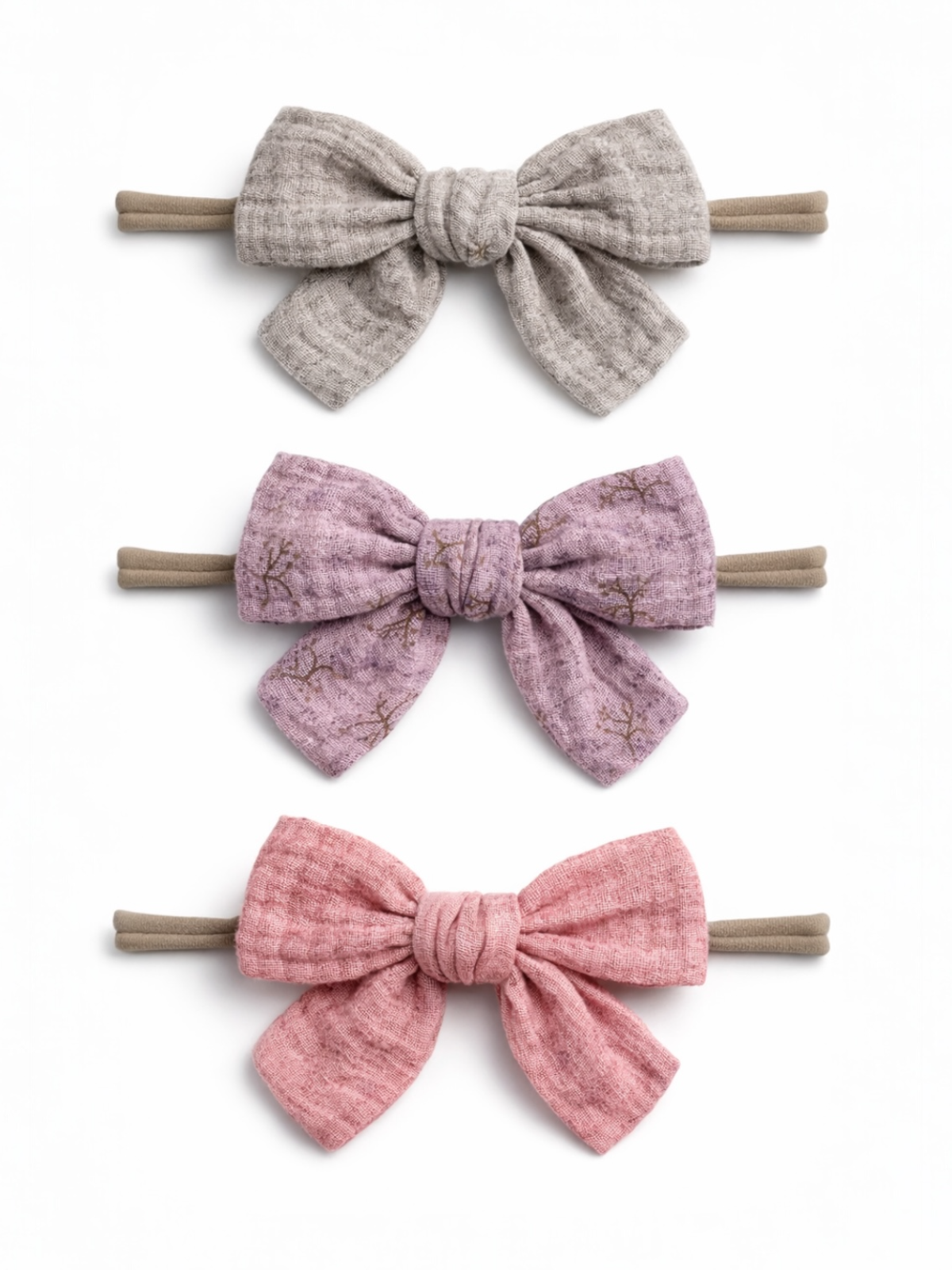 Three fabric hair bows in gray, lavender, and pink, each secured on elastic bands, displayed against a white background.