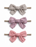 Three fabric hair bows in gray, lavender, and pink, each secured on elastic bands, displayed against a white background.