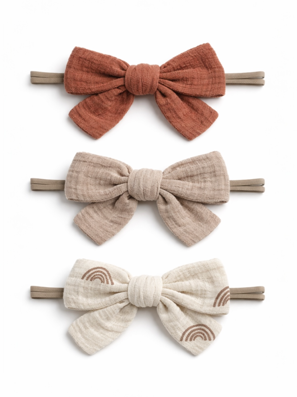 Three fabric hair bows in rust, beige, and cream with rainbow patterns, displayed on elastic bands.