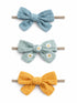 Three colorful fabric hair bows: blue, green with daisies, and yellow, tied on elastic bands.