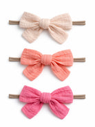 Three fabric hair bows in soft colors: cream, coral, and pink, attached to elastic bands.