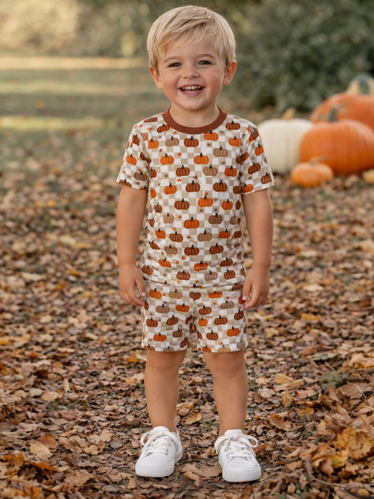 Smiling toddler in pumpkin-patterned outfit standing on fallen leaves, surrounded by pumpkins in a vibrant autumn setting.